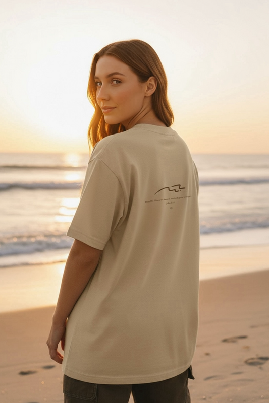 Side view of a woman wearing a beige “Grace flows like the ocean” tee while standing by the beach.