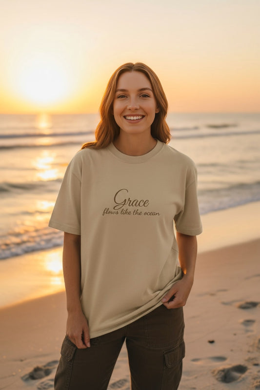 Woman walking on the beach wearing the beige “Grace flows like the ocean” oversize tee.