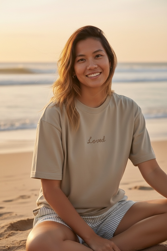 Woman sitting on a beach wearing a beige t-shirt with 'Loved' text.