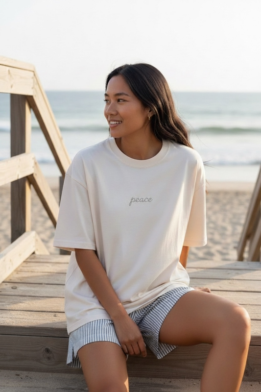 Woman sitting on a wooden bench by the beach wearing a white 'peace' t-shirt.